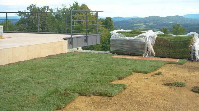 Two unpacked stacks of grass patches lie next to a balcony and unfinished backyard of a big housing project. The yard of a modern house in countryside gets renovated with patches of artificial grass.