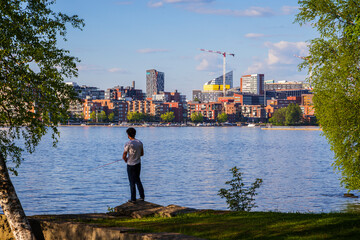 Man fishing at the Hatanpää arboretum, view of Tampere city over the lake Pyhäjärvi in Finland in the summer. - obrazy, fototapety, plakaty
