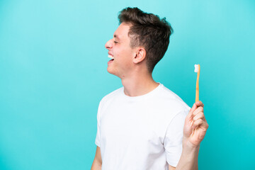 Young Brazilian man brushing teeth isolated on blue background laughing in lateral position - obrazy, fototapety, plakaty