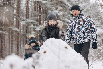 Happy father and sons rolling big snowball together for snowman in snowy winter forest. Man Joyful...