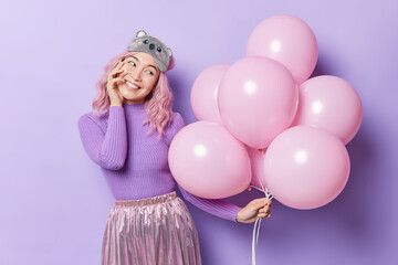 Obraz na płótnie Canvas Horizontal shot of pretty young Asian woman holds bunch of inflated balloons prepares for party or celebration wears casual jumper skirt and sleepmask on forehead isolated over purple background