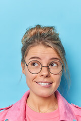 Vertical shot of beautiful young woman concentrated above with dreamy expression bites lips considers something wears round spectacles isolated over blue background. People and thoughts concept