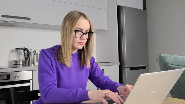 Pensive young adult woman in glasses work study at kitchen using laptop. Work from home concept