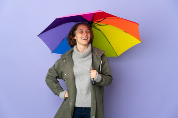 English woman holding an umbrella isolated on purple background posing with arms at hip and smiling