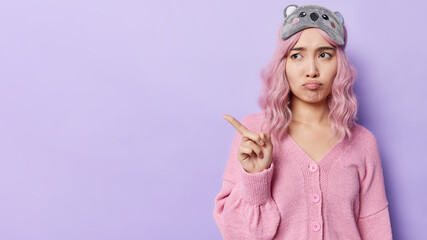 Horizontal shot of dissatisfied sulking Asian woman points away at blank space doesnt like something feels upset wears sleepmask and casual jumper isolated over purple background. Look at this