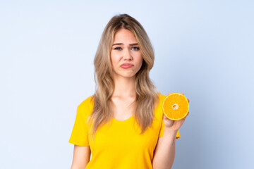 Teenager Russian girl holding an orange isolated on blue background with sad expression