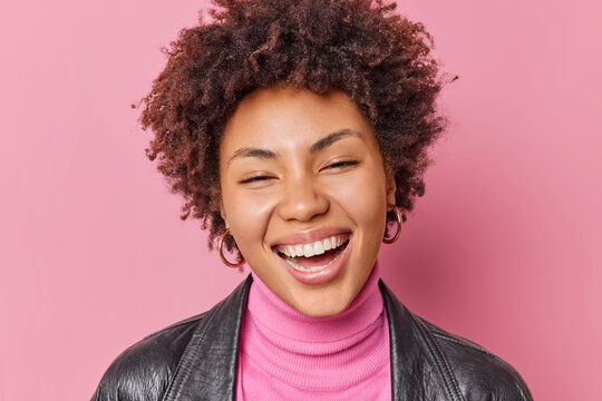 Portrait of good looking woman with broad toothy smile expresses happy emotions feels glad shows white teeth wears turtleneck and leather jacket isolated over pink background. Happiness concept