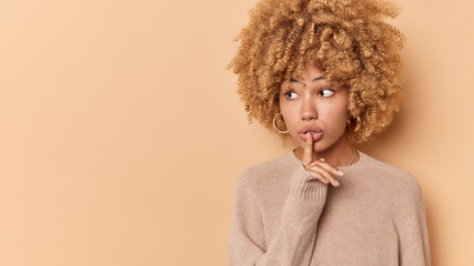 Keep mouth shut. Mysterious curly haired woman makes silence gesture holds index finger over lips gossips or spreads rumors wears casual jumper isolated over beige background blank copy space