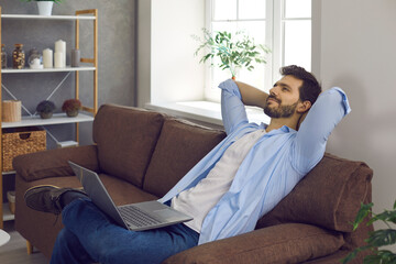 Obraz premium Happy young man takes break from work or studies. Relaxed handsome entrepreneur or college student lounging on comfortable sofa with laptop computer on lap, thinking hands behind head, daydreaming