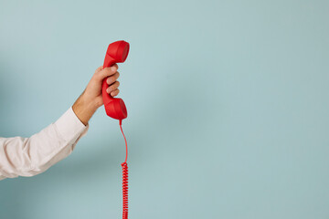 Unidentified man holding red wired retro telephone handset on light blue banner background. Male hand holds handset of landline phone aimed at free space for text. Customer support concept. - obrazy, fototapety, plakaty