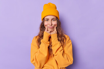 Upset young woman with long hair holds fingers at edge of lips forces smile has unhappy expression...