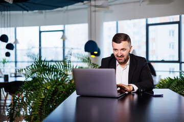 Portrait of a stylish young business man, in a large modern office high on an upper floor, looking into a laptop. Business man at freelancer finance office desk