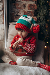 little boy in an elf hat with a candy in his hand sitting at the window. Child looks out the window and waits for Christmas, Santa Claus