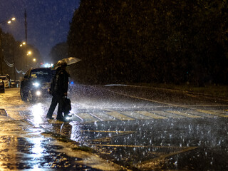 View of a pedestrian crossing in the city at night during a heavy downpour. Silhouettes of people with umbrellas in the headlights of the car. People cross the road. 