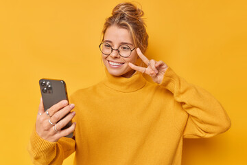 Positive young woman makes peace gesture takes selfie makes photo of herself dressed in casual...