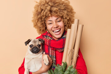 Portrait of happy curly haired woman pet owner poses with pedigree dog and new year decorations smiles ladfully has positive festive mood dressed in winter clothes isolated over brown background