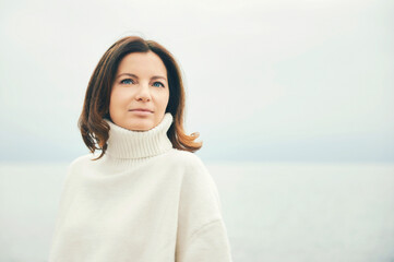 Outdoor portraiit of beautiful woman wearing white pullover, relaxing next to lake or sea
