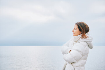 Outdoor portraiit of beautiful woman wearing white jacket, relaxing next to lake or sea