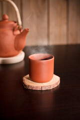 Earthenware mug with hot steam, coffee or tea on wooden table, with partial view of kettle, brown wooden background.