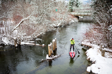 A paddle boarder paddles up a Canadian river during a snowstorm.  Whistler BC, Canada.