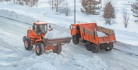 Big orange tractor cleans up snow from the road and loads it into the truck. Cleaning and cleaning...