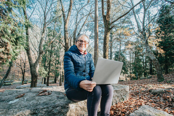 Happy mature woman having a video chat through laptop computer while a day at the forest during a travel to treck. Copy space sitting on a wood bench. Active senior woman healthy life style.