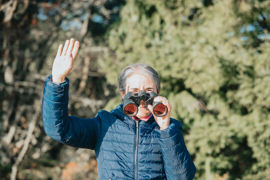 Beautiful Senior Woman Using Binoculars To Spot Animals And Things While Hiking With Backpacks And Trekking In The Forest. Concept Of Active Lifestyle On Retirement. Senior People New Habits New Year