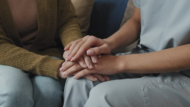 Close up of unrecognizable senior woman and nurse sitting with holding hands on sofa
