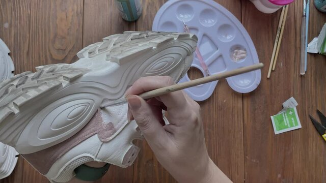 Female's hands dip brush in brown paint and apply it to sneaker. Palette, tools and tubes on wooden table. Hobbies, manual work and leisure at home. Design of sports shoes using acrylic.