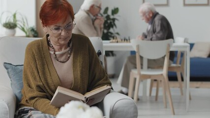 Medium slowmo shot of red-haired Caucasian senior woman reading book while two senior men playing chess in background, living together in nursing home - Powered by Adobe