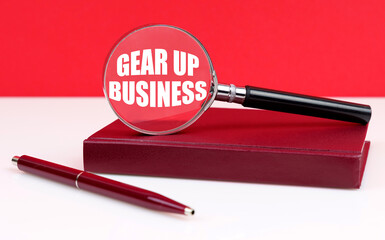 On a white and red background, a notebook, a pen and a magnifying glass, inside which the inscription - Gear Up Business