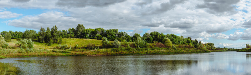 Wide panoramic view of calm lake and green hills. Grey stormy clouds in dramatic sky.Summer natural landscape.
