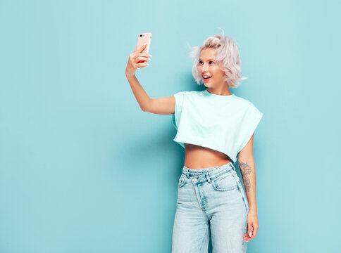 Young beautiful smiling female in trendy summer clothes. Sexy carefree woman posing near blue wall in studio. Positive blond model having fun and going crazy. Cheerful and happy.Taking selfie photos