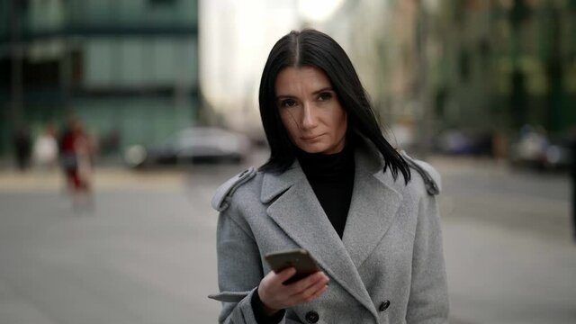 portrait of a brunette in a gray coat and with a phone in hand against the background of a blurry modern urban area. the camera is moving