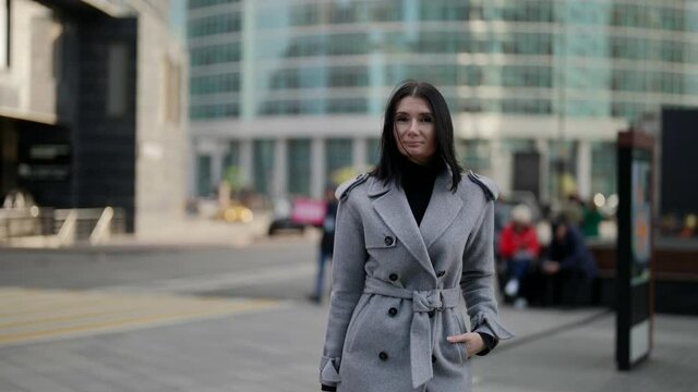 a brunette in a gray coat has hand in pocket and is walking against the background of modern buildings. the middle plan