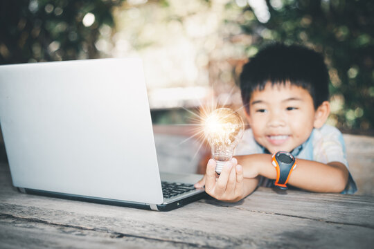 Asian boy with a smiling face using a notebook computer in the hand holding a glowing light bulb represents modern technology to use the internet for learning in the wider world.