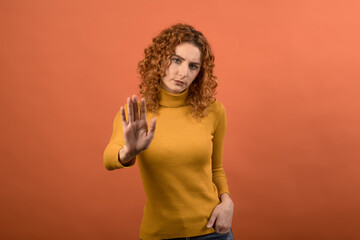 Young and attractive redhead Caucasian girl in orange jumper extending palm and showing STOP gesture isolated on orange studio background.