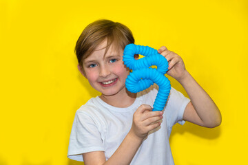 happy schoolboy in a white t-shirt holds a blue corrugated pop tube зantistress toy. smiling child stands on yellow background