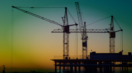 Construction cranes are building a house against the background of sunset and dark sky