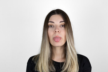Fototapeta premium Young woman sticking out tounge, close up portrait. Teasing you through camera while isolated with white background. 