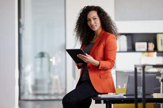 Smiling businesswoman holding tablet PC while sitting on desk in office
