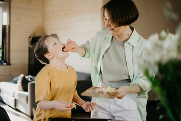 Smiling mother feeding grated cheese to son covered with flour on face at home