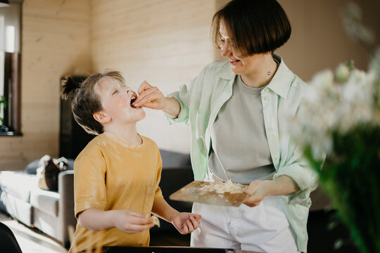 Smiling mother feeding grated cheese to son covered with flour on face at home