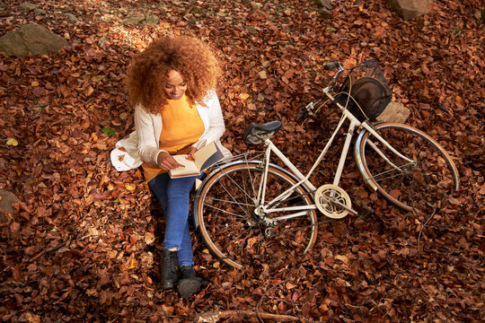 Woman with bicycle reading book sitting on fallen leaves in autumn forest