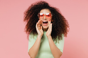 Promoter young curly latin woman 20s years old wears mint t-shirt sunglasses scream hot news about sales discount with hands near mouth isolated on plain pastel light pink background studio portrait. - obrazy, fototapety, plakaty