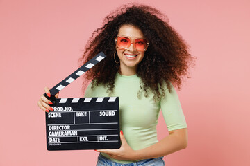 Exultant happy charismatic vivid young curly latin woman 20s wear mint t-shirt sunglasses holding classic black film making clapperboard isolated on plain pastel light pink background studio portrait - obrazy, fototapety, plakaty