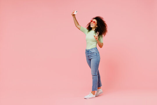 Full size happy fancy young curly latin woman 20s wears casual clothes do selfie shot on mobile cell phone post photo on social network isolated on plain pastel light pink background studio portrait.