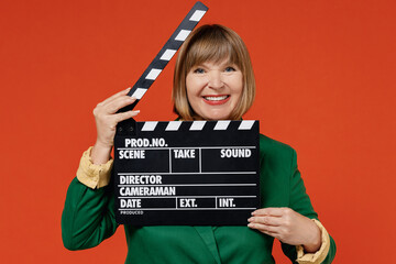 Elderly smiling fun woman 50s wearing green classic suit holding classic black film making clapperboard isolated on plain orange color background studio portrait. People business lifestyle concept