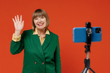 Elderly smiling happy woman 50s wear green classic suit talk by video call using mobile cell phone on stand waving hand isolated on plain orange color background. People business lifestyle concept