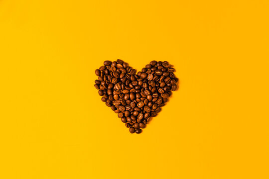 A stylish heart made of coffee beans on a yellow background. Freshly roasted Arabica seeds on a light background with space for text for your advertising